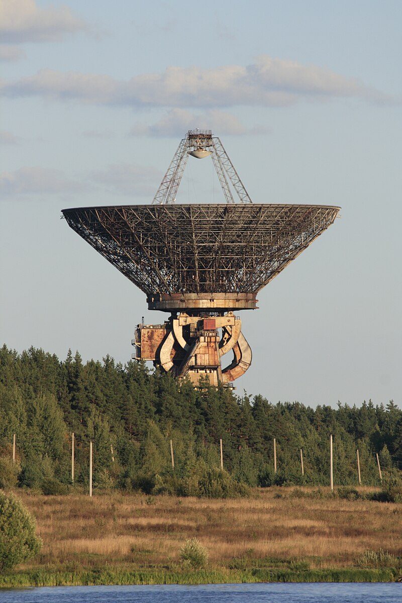 Large, rusted metal radio telescope dish with lattice structure rising above a dense forest near a body of water under a partly cloudy sky.