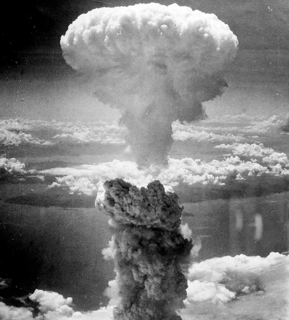 Black and white photograph of a large mushroom cloud rising above scattered clouds, showing a dense vertical column of smoke and debris billowing outward near the top.