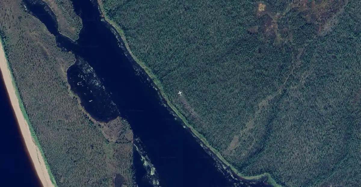 Aerial view of a dense forest area adjacent to a winding river, with a small white airplane wreckage visible near the riverbank amidst the trees.