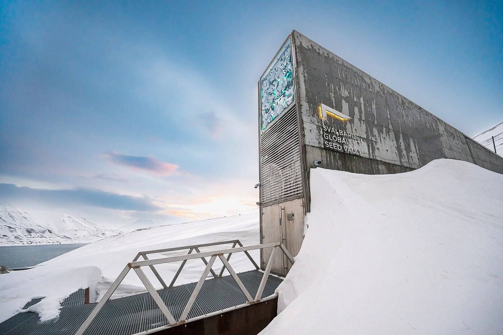 Snow-covered entrance of the Svalbard Global Seed Vault built into a mountainside with a metal bridge leading to a heavy door, surrounded by snowy Arctic landscape under a partly cloudy sky at sunset.